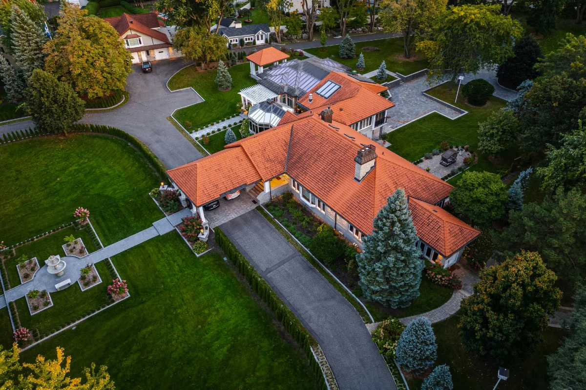 Aerial view of a large house with red-tile roof, manicured lawn, and winding driveway surrounded by trees and gardens in a suburban estate.