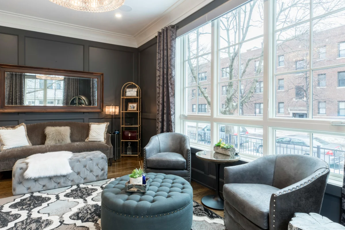 Living room with dark paneled walls, a large window, gray upholstered chairs, a tufted blue ottoman, and a patterned rug under a coffee table centerpiece.