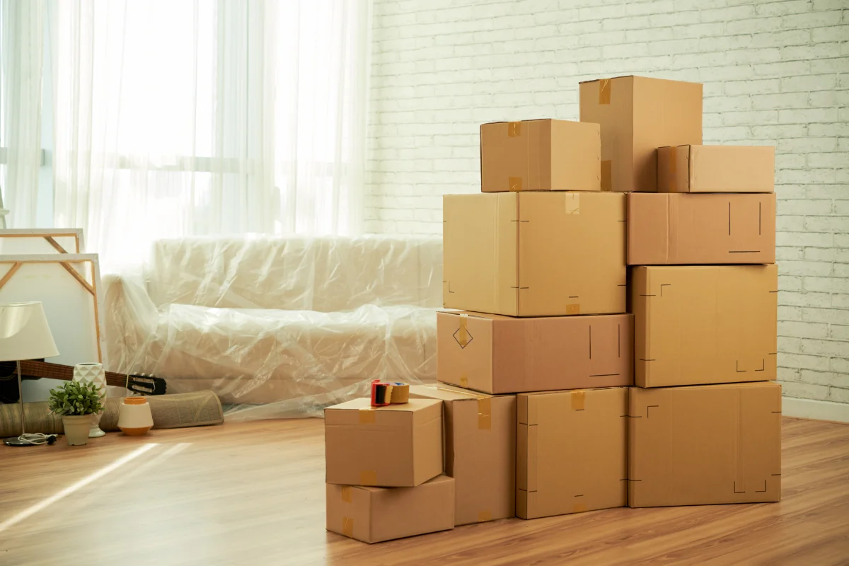 Stack of taped cardboard moving boxes arranged in a bright living room with a white brick wall and wooden floor