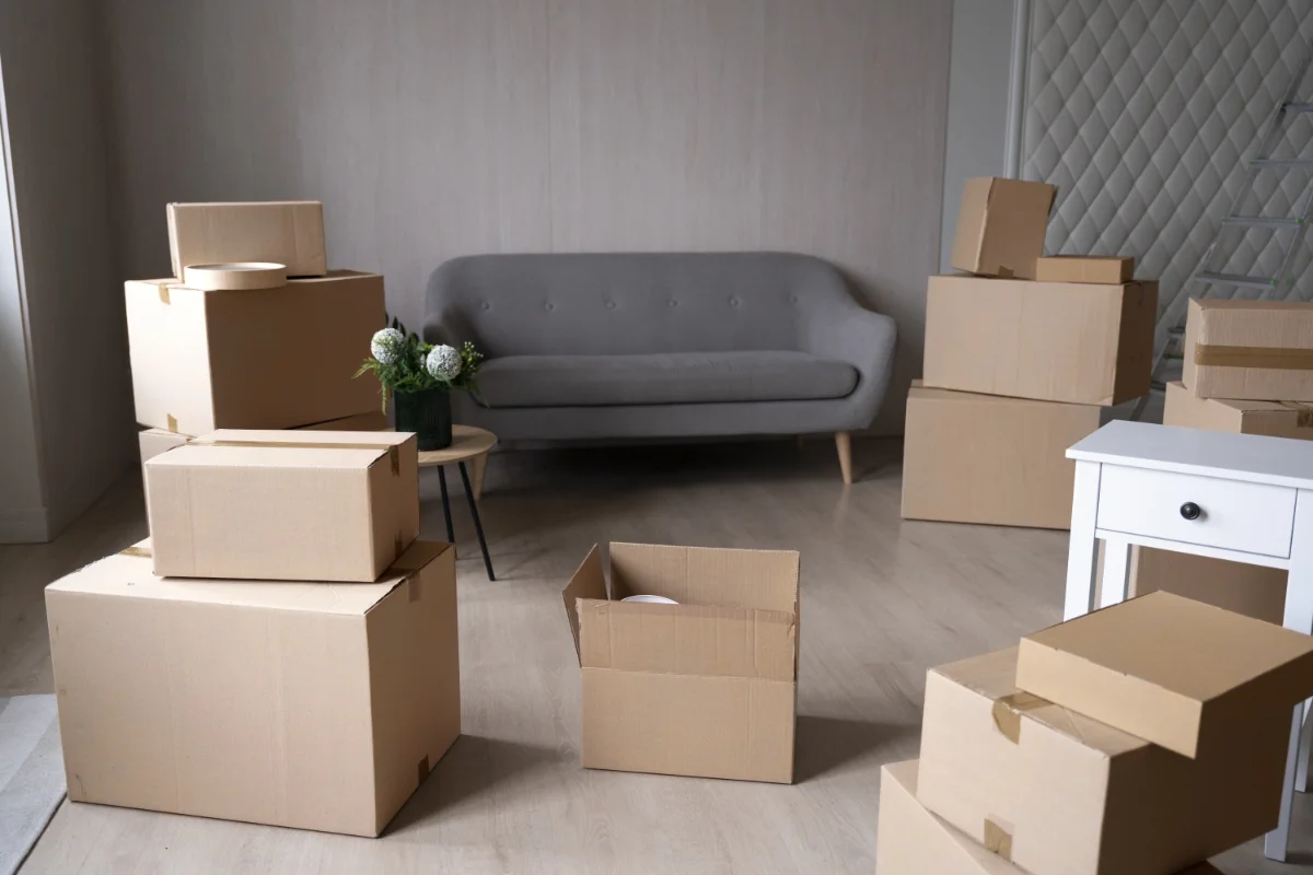 Living room with stacked moving boxes around a gray sofa and a white desk in the background in a home during a move.