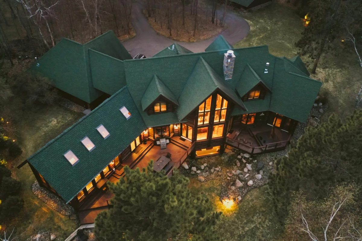 A large green-roofed cabin-style house at dusk, with warm interior lights visible through many windows, surrounded by trees.