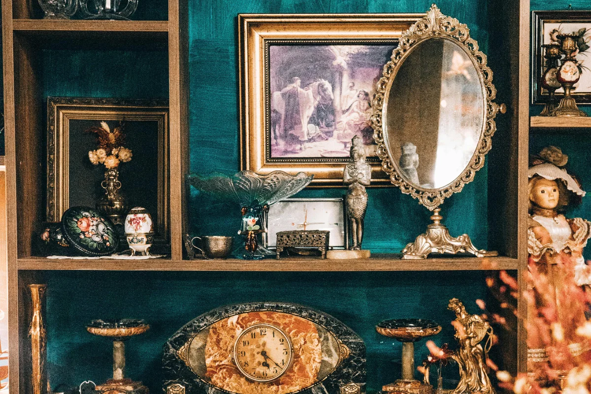 Eclectic vintage shelf with antique clock, framed artwork, ornate mirror, and porcelain dolls.