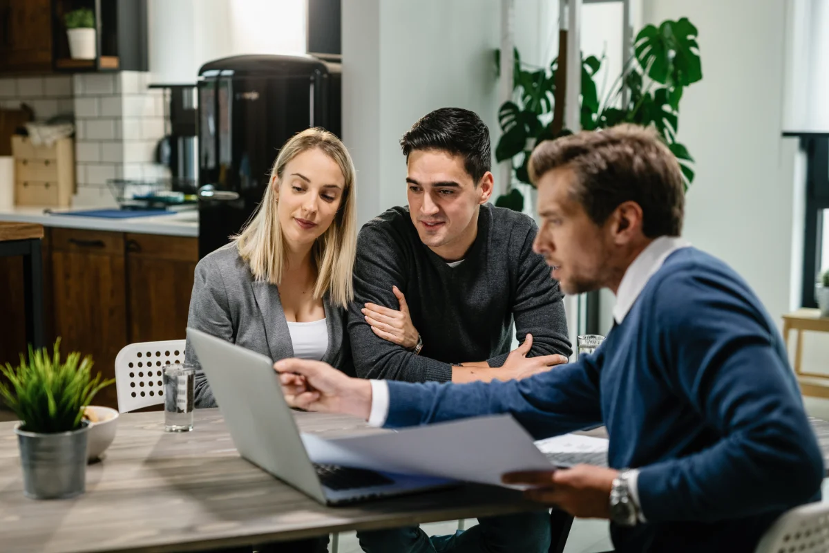 Three colleagues sit at a wooden table in a modern office kitchen, reviewing a laptop and documents together as they discuss a project.