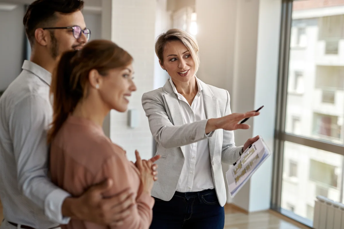 A woman in a light blazer presents a document to two colleagues in a bright office, gesturing with a pen as they listen nearby.